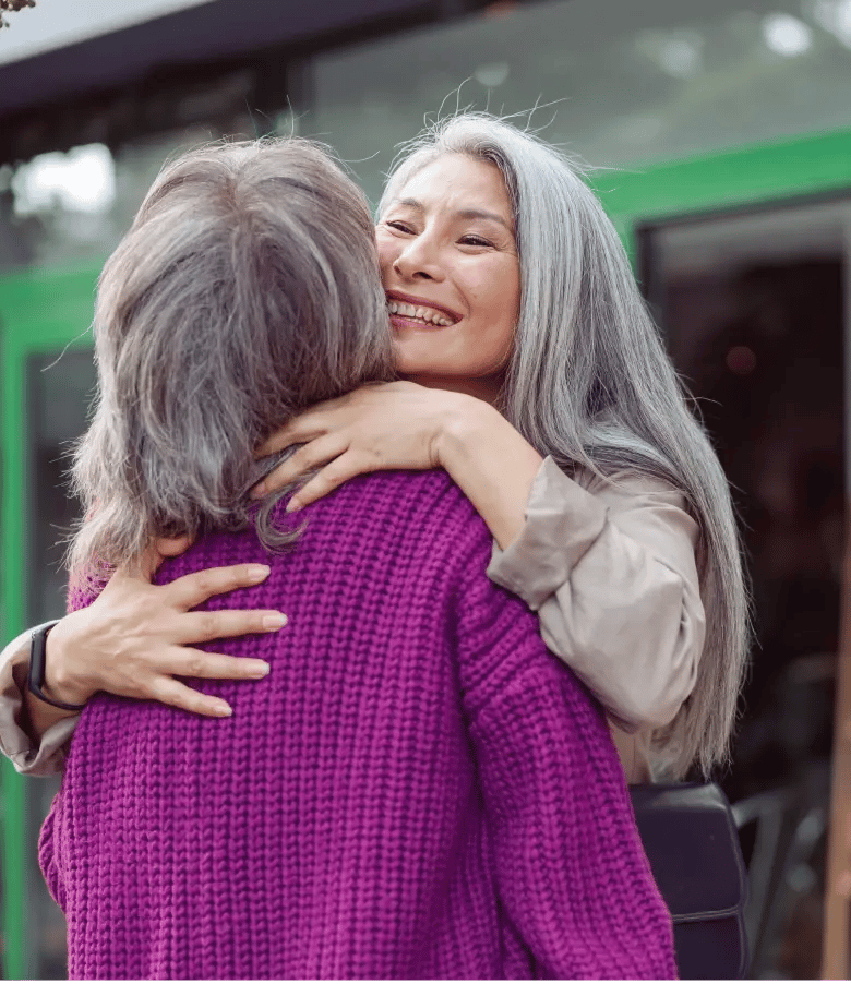 Two women with long gray hair embrace warmly outside a café, one wearing a purple sweater and the other in a beige top.